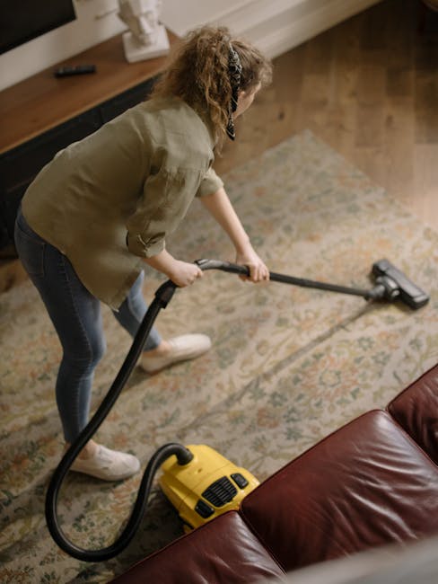 Close-up view of a vacuum cleaner with a transparent dust container filled with debris, connected to a flexible, patterned hose and a clean black floor attachment, positioned on a beige carpet in a ro
