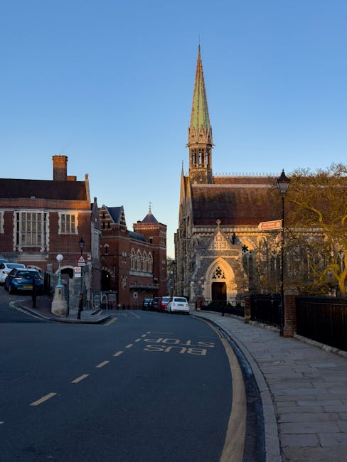 A narrow street in Ealing with a bus lane marked on the asphalt and parked cars on the side. The street is lined with traditional red brick buildings, including a church with a tall, pointed spire and