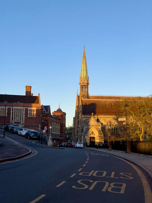 A narrow street in Ealing with a bus lane marked on the asphalt and parked cars on the side. The street is lined with traditional red brick buildings, including a church with a tall, pointed spire and